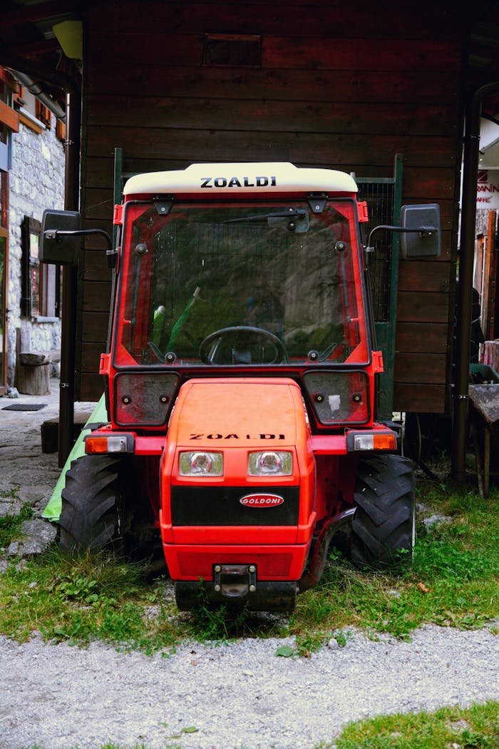 Bright red tractor parked between rustic farm buildings in a rural setting.